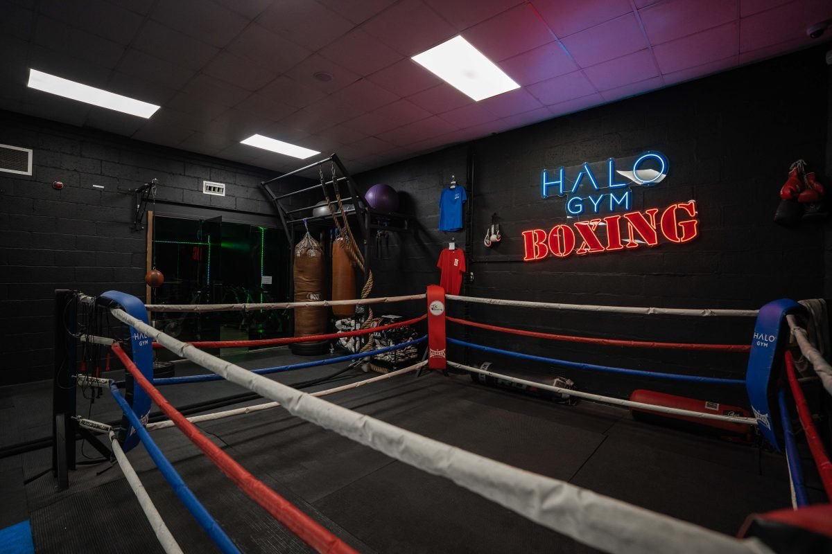 Boxing ring at Halo Gym with red, white, and blue ropes, punching bags, and a neon Halo Gym Boxing sign on the wall.